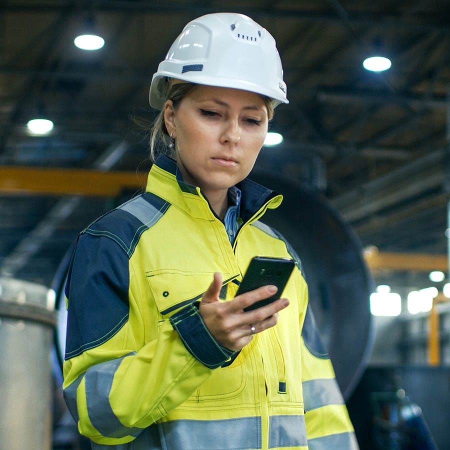Female worker in safety gear using a smartphone in an industrial setting, illustrating micro-learning in the workplace.